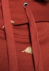 Rød hættetrøje med tekstureret stof, gyldent accentmønster og tykke, fletrede snore med sølvfarvet øje.