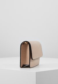 Beige texturovaná kabelka přes rameno s černými detaily, vystavená vzpřímeně na bílé ploše na světle šedém pozadí.