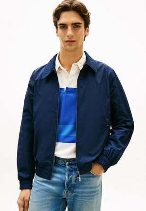 Ung mand med brunt hår iført en marineblå lynlåstrøje, hvid og blåstribet polo, og lyseblå jeans, stående med den ene hånd i lommen.