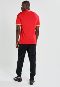 Maglietta atletica rossa con accenti gialli, abbinata a pantaloni neri e scarpe da calcio. La maglietta ha maniche corte e un collo a giro.