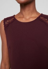 Bordowa, bezrękawnikowa bluzka z misternymi koronkowymi detalami na ramionach oraz gładką, miękką fakturą, z okrągłym dekoltem.