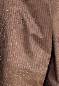 Tissu imperméable marron avec un motif texturé ; gouttes d'eau sur la surface ; variation de couleur distincte au niveau de la couture.