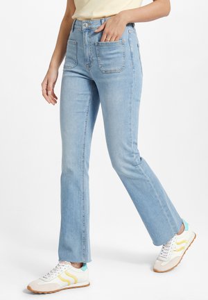 Persoon draagt lichtblauwe rechte jeans met opgestikte voorzakken en witte sneakers met gele en blauwe accenten.