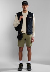 Chaleco de marina con múltiples bolsillos sobre un suéter beige, pantalones cortos oliva, zapatos negros y una gorra a juego, sosteniendo una cantimplora negra.
