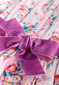 Gėlėta suknelė, pagaminta iš rožinės medžiagos su ryškiais gėlių raštais. Didelė purpurinė aksominė juosta puošiama ties juosmeniu, pabrėždama dizainą.