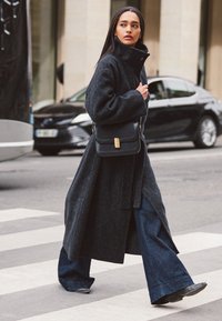 Long dark gray wool coat with high collar, paired with wide-leg denim trousers, and a black handbag featuring a gold clasp. Urban street setting.