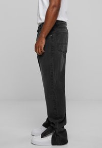 Black denim jeans with a relaxed fit, visible side zippers at the ankles, and back patch pockets. Paired with white sneakers.