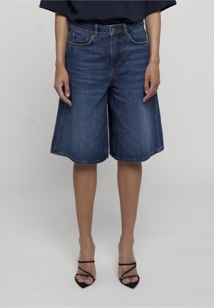 Person iført mørkeblå denim knælange shorts og sorte stroppede højhælede sko, der står mod en ensfarvet baggrund.
