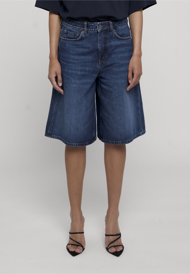 Personne portant un short en jean bleu foncé longueur genou et des chaussures à talons hauts noires à brides, debout devant un fond uni.