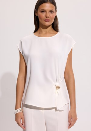 Kobieta w białej, bezrękawnikowej bluzce z bocznym węzłem i złotymi guzikami, w połączeniu z białymi spodniami i perłową bransoletką.