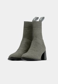 Un par de botas de tobillo de tela acanalada en verde oliva con tacones cuadrados y tiradores etiquetados "CAMPER" sobre un fondo blanco.