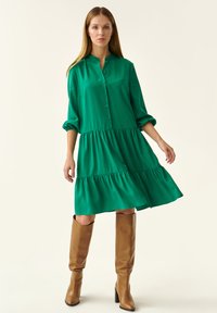 Vestido verde con botones y diseño en capas. Manga larga y corte relajado. Combinado con botas altas marrones. Tela suave y hasta la rodilla.