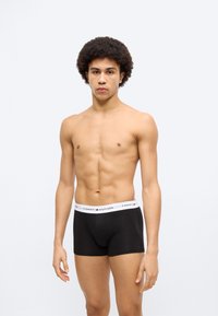 Μαύρα ανδρικά εσώρουχα boxer με λευκή μέση που φέρει το λογότυπο "TOMMY HILFIGER", φορεμένα από άνδρα μοντέλο με σγουρά, κοντά μαλλιά.