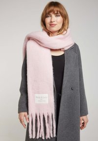 Écharpe en laine rose clair avec des bords frangés, arborant une étiquette "Andy & Lucy" et une texture douce, portée sur un manteau gris foncé.
