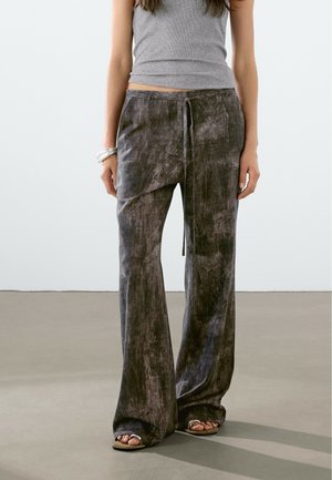 Femme portant un pantalon large en velours gris foncé avec un nœud à l'avant et un haut gris sans manches, debout sur un sol lisse gris clair.