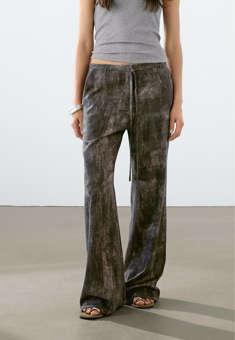 Femme portant un pantalon large en velours gris foncé avec un nœud à l'avant et un haut gris sans manches, debout sur un sol lisse gris clair.