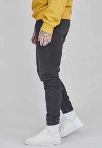 Schwarze, schmal zulaufende Jogginghose mit elastischen Bündchen, kombiniert mit weißen Turnschuhen. Das Outfit umfasst einen gelben Sweatshirt und sichtbare Tattoos an der Hand.