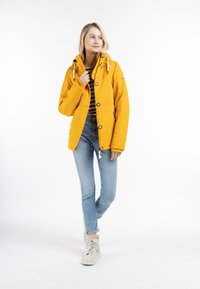Chubasquero amarillo impermeable con capucha, que presenta dos grandes botones, detalles con cordones y dos bolsillos laterales, combinado con jeans de un azul claro.