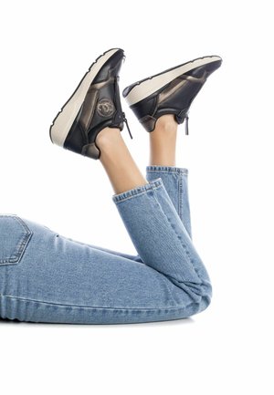 Zapatos deportivos negros y de bronce con un diseño texturizado, estilo slip-on y suelas gruesas blancas, usados con jeans ajustados de color azul claro.