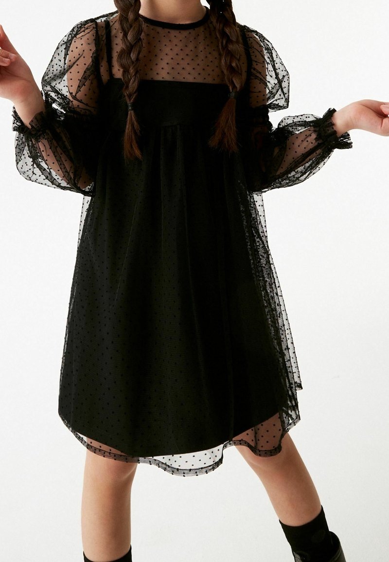 Schwarzes Kleid aus transparenter Pünktchenstoff, mit puffartigen Ärmeln und einem quadratischen Ausschnitt, verziert mit Rüschenakzenten an den Ärmelenden.