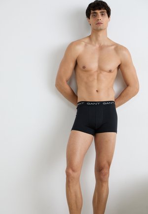Schwarze Herren-Boxershorts mit Logobund, aus weichem Stoff, figurbetontem Schnitt und mittellanger Beinform, auf einem nackten Oberkörper vor einer weißen Wand.