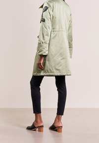 Giacca verde chiaro, fino al ginocchio, con una finitura testurizzata e toppe ricamate a forma di foglia, abbinata a pantaloni neri aderenti e zoccoli con tacco in legno.