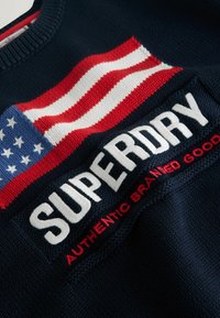 Jūras zilas trikotāžas sporta krekls ar Amerikas karoga dizainu un izteiktu baltu "SUPERDRY" logo ar sarkaniem akcentiem zem tā.