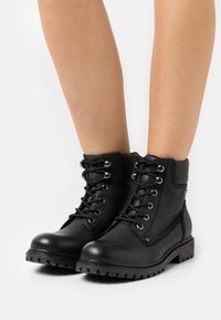 Gambe che indossano stivali alla caviglia in pelle nera con lacci e suole robuste, mostrati su uno sfondo bianco semplice.