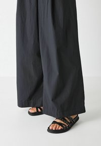 Black sandals made of rubber with multiple straps and stud accents. Worn with loose-fitting black fabric trousers.