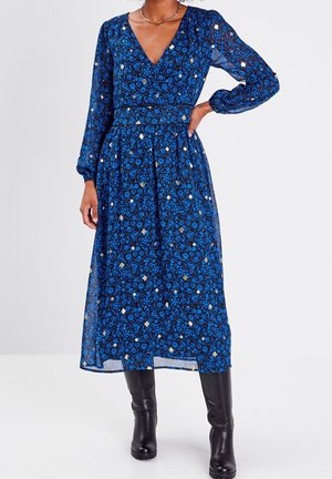 Robe longue à manches longues à fleurs bleues avec un décolleté en V profond, taille froncée et tissu transparent. Elle présente des motifs géométriques dorés et blancs, associée à des bottes noires.