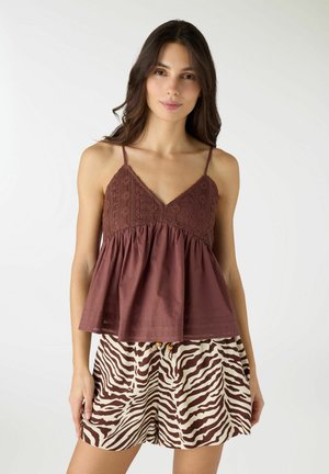 Donna che indossa una canottiera marrone con corpetto in pizzo e pantaloncini a stampa zebra, in piedi davanti a uno sfondo chiaro e uniforme.