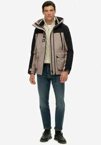 Beige en zwarte jas met ritszakken, trekkoorden en logodetail, gecombineerd met een lichte trui en blauwe jeans. Zwarte schoenen.