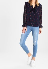 Donna che indossa una blusa trasparente blu navy con stampe floreali rosse, jeans skinny chiari strappati e sneakers bianche, in piedi contro uno sfondo bianco.