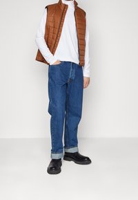 Braune gesteppte Weste über einem weißen Langarmshirt, kombiniert mit blauen Jeans und schwarzen Schuhen. Einfaches Design mit einem Frontreißverschluss.