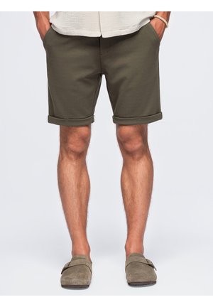 Homme portant un short vert olive à ourlet roulé, les mains dans les poches et des chaussures beige sans lacets, debout devant un fond uni.