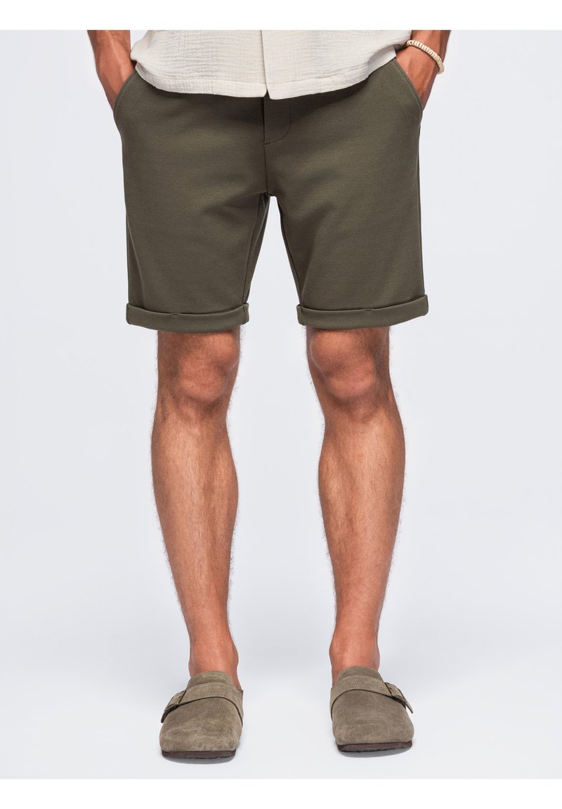 Homme portant un short vert olive à ourlet roulé, les mains dans les poches et des chaussures beige sans lacets, debout devant un fond uni.
