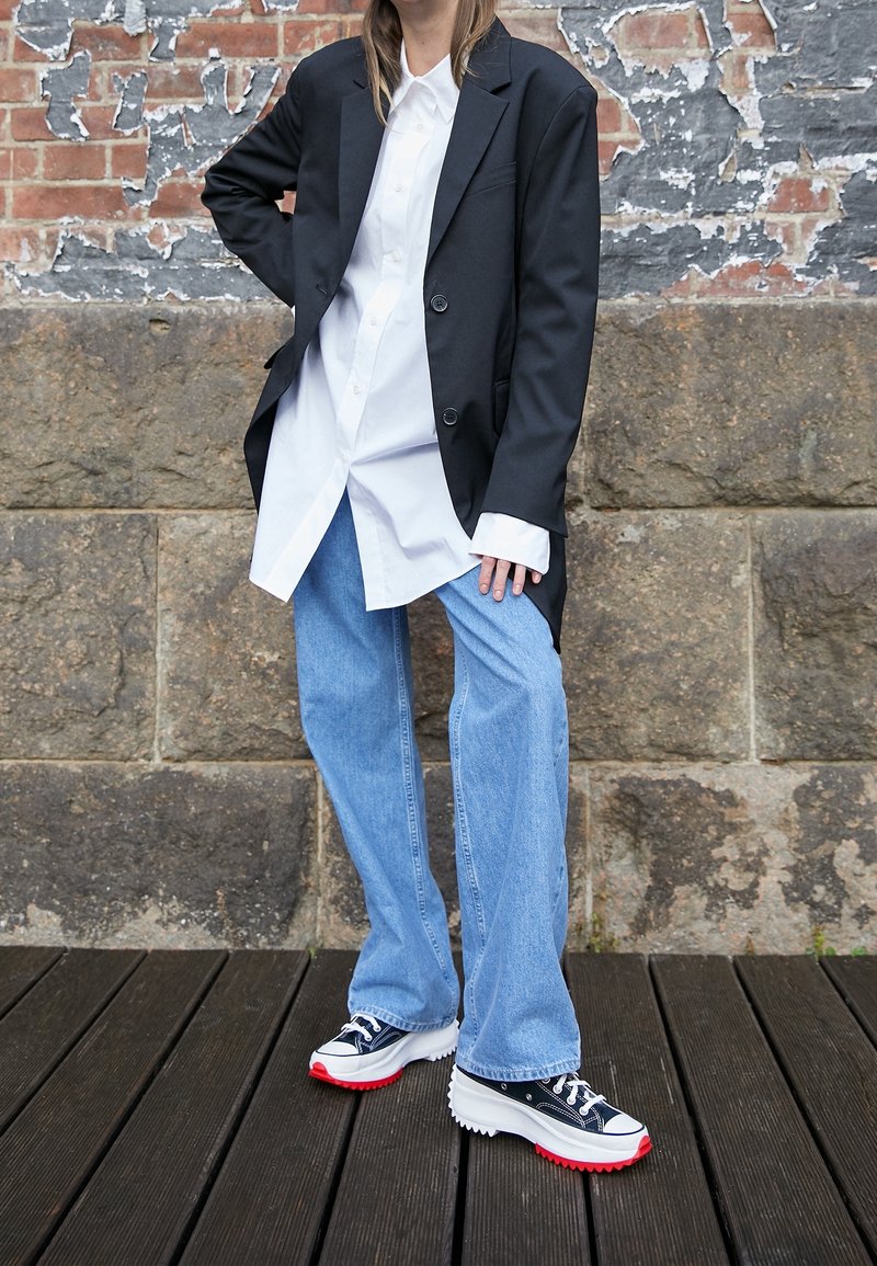 Personne portant un blazer noir oversize, une longue chemise blanche, un jean large bleu clair et des baskets plateformes noires avec des semelles rouges, debout contre un mur en briques.