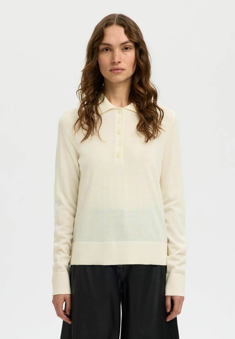 Femme aux longs cheveux ondulés portant un polo crème à manches longues et un pantalon noir, debout devant un fond blanc uni.