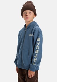 Blauwe hoodie met rits, voorzien van voorzakken en grafische tekst op de mouwen. Gepaarde met een bruine beanie. Stof lijkt zacht en comfortabel te zijn.