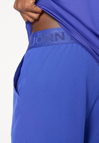 Calças de desporto azuis com uma textura suave, apresentando uma cintura elástica larga com a inscrição "JÖRN" e bolsos laterais para maior funcionalidade.