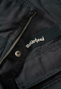 Juodas audinio švarkas viduje su sidabriniu užtrauktuku ir tamsiu padu, ant kurio metaliniais raidėmis parašyta "Motörhead". Ryškūs siuvimo akcentai.