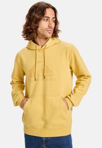 Sudadera con capucha amarilla en tela suave, con un bolsillo tipo canguro en la parte frontal, capucha con cordones y logotipo en relieve en el pecho.