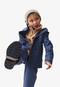 Marine blauwe waterdichte jas met een rits, beige fleece voering en bijpassende broek. Voorzien van een gebreide muts, verhoogde kraag en een gestructureerde zool.