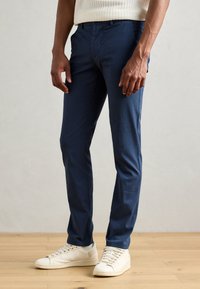 Chinos bleu marine en tissu lisse, avec une coupe slim et un devant plat. Associés à des baskets blanches sur un sol en bois.
