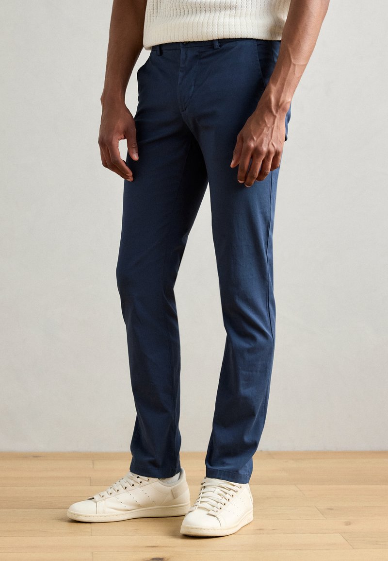 Chinos bleu marine en tissu lisse, avec une coupe slim et un devant plat. Associés à des baskets blanches sur un sol en bois.