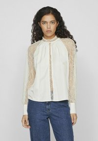Blusa blanca con mangas largas de encaje, cuello alto y cierre frontal con botones. El cuerpo es de una tela sólida, en contraste con las mangas transparentes.