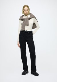 Čierne nohavice s rovný strihom, krémová košeľa s dlhým rukávom a svetlohnedý pletený sveter prehodený cez ramená. Čierne topánky.