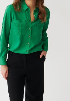 Femme portant une chemise boutonnée vert vif avec poches avant et pantalon noir, main dans la poche, sur un fond clair uni.