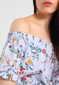 Blůza bez ramínek ve světle modré s bílými pruhy, s květinovým vzorem v různých barvách. Texturovaná tkanina se nařaseným výstřihem.