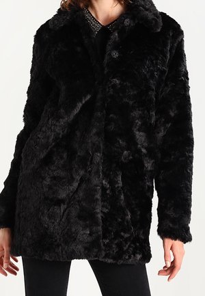 Manteau en fausse fourrure noire avec une surface texturée douce, fermeture à boutons, porté sur un haut foncé et un pantalon noir.